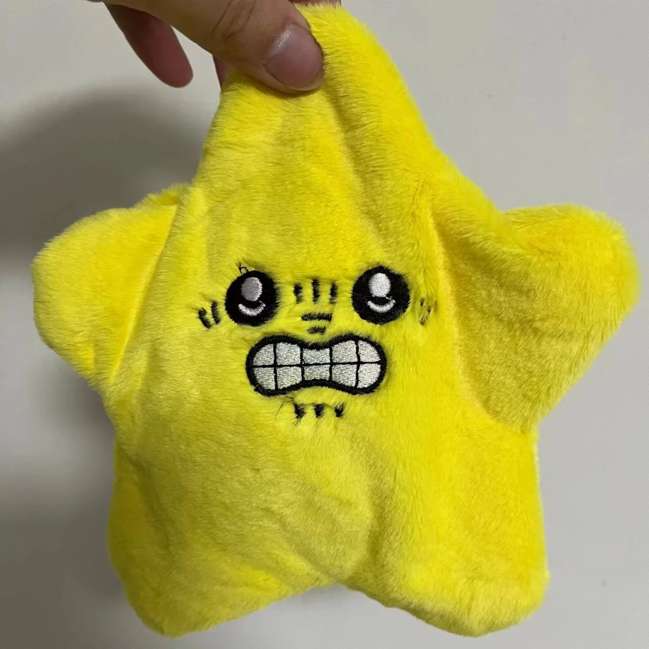 Plush yellow star toy with an embroidered silly face, held by a hand, on plain background